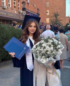 a woman in graduation gown holding a bouquet of flowers and a blue folder on the street