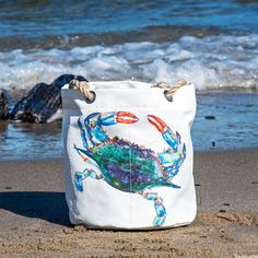 a white bag sitting on top of a sandy beach next to the ocean with blue crabs painted on it