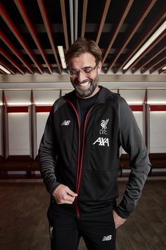 a man with glasses and a beard wearing a black liverpool football hoodie smiles at the camera