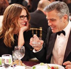 two people sitting at a table with wine glasses in front of them and one person wearing a tuxedo