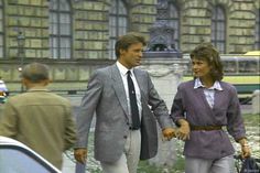 the man and woman are walking together in front of a fountain with cars behind them