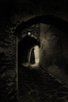 a dark tunnel with a person standing in the light at the end and a lantern on the wall