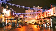 an empty street is lit up at night with lights strung from the buildings and tables