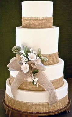 a three tiered wedding cake with burlap ribbon and flowers on top, sitting on a table