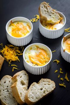 four small white bowls filled with eggs and bread on top of a table next to slices of bread