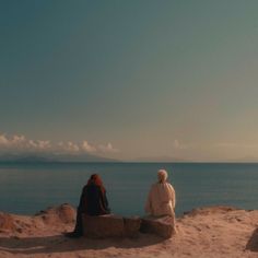 two people sitting on top of a sandy beach next to the ocean and looking out at the water