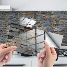 two hands holding up a piece of glass in front of a stone wall and stove