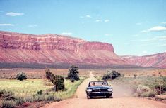 an old car driving down a dirt road in front of red rocks and green grass