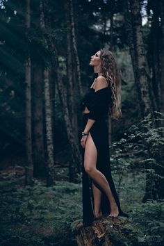 a woman standing on top of a tree stump in the woods wearing a black dress