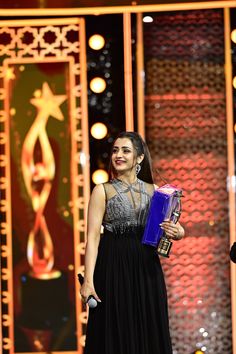 a woman in a long black dress holding an award on stage with lights behind her