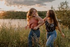 two young women are walking in tall grass near the water at sunset or sunrise,