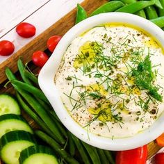 a white bowl filled with dip surrounded by green beans, cucumbers and tomatoes