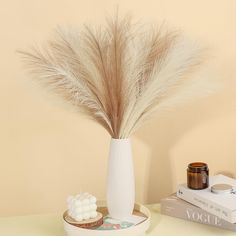 a white vase with some dry grass in it on a table next to a book