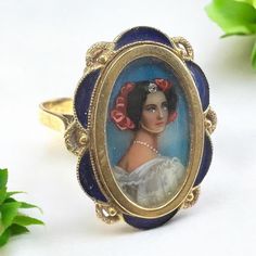 This handpainted Victorian Minature Portrait Ring from the 1800s has a lovely woman with a white dress and what appears to be red flowers in her hair. In the center of her forehead is an actual tiny diamond. Pearls are painted around her neck. The painting is set into a scalloped frame with blue enamel which is just the right accent for the artwork.  18 karat yellow gold Stamped "750" Size 8 1/4 - can be sized  Measurements: 7/8" x 5/8"  Weight: 3.2 Dwt. (4.9 Grams) Victorian Enamel Ring For Anniversary, Victorian Hallmarked Enamel Ring For Wedding, Victorian Style Hallmarked Enamel Ring For Wedding, Antique Hallmarked Enamel Wedding Ring, Victorian Yellow Gold Enamel Ring, Antique Enamel Wedding Ring, Antique Gold Enamel Wedding Ring, Antique Yellow Gold Enamel Wedding Ring, Antique Gold Enamel Ring For Wedding