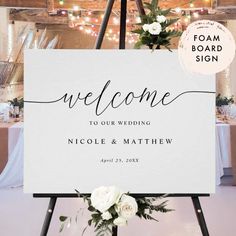 a welcome sign with white flowers on it in front of a wooden table and lights