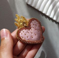 a hand holding a pink heart shaped brooch with gold angel wings on it's back