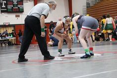 two women wrestling in a gym with an older man watching from the sidelines behind them