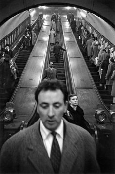 a man wearing a suit and tie standing in front of an escalator filled with people