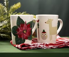 two coffee mugs decorated with christmas designs on a red and white checkered table cloth