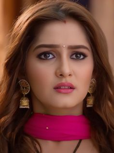 a woman with long brown hair wearing large earrings and a pink dress is looking at the camera