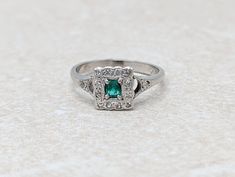 This captivating cluster ring centres on an emerald cut rectangular emerald, surrounded by a cluster of diamonds, complemented by a diamond to each shoulder. The scalloped edging exudes a timeless elegance and charm. Ring size: 4.25 (re-sizing available for additional fee - please enquire) Condition: Excellent, commensurate with age Era circa: Mid century Stamps: 18k gold Approx. measurements: 7mm full head width, 3mm full head height Stones: 1 x emerald estimated 0.15 carat weight, 14 x diamond Rectangular Emerald Diamond Ring For Formal Occasions, Formal Rectangular Emerald Ring With Center Stone, Formal Rectangular Emerald Diamond Ring, Emerald Cluster Ring In White Gold, Rectangular Emerald Ring With Brilliant Cut Diamond, Art Deco Rectangular Emerald Ring For Anniversary, Rectangular Emerald Ring With Center Stone For Wedding, Formal Emerald Cut Ring With Single Cut Diamonds, Art Deco Rectangular Emerald Anniversary Ring