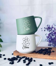 two coffee mugs sitting on top of each other next to some black beans and purple flowers