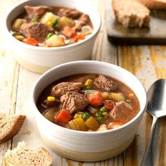 two bowls of beef and vegetable soup on a wooden table next to slices of bread