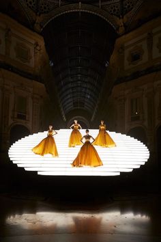 three women in yellow dresses are dancing on a large white surface with lights behind them