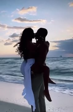 a man holding a woman on the beach at sunset with waves crashing in front of him