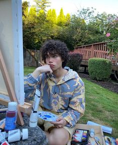 a young man sitting on the ground next to an easel filled with paint and supplies