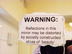 a black and white photo of a sign warning people to reflect in this mirror may be distorted by socially constructed ideas of beauty