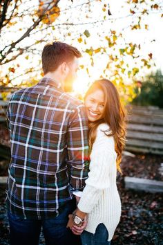 an engaged couple holding hands and smiling at each other in front of a tree with leaves