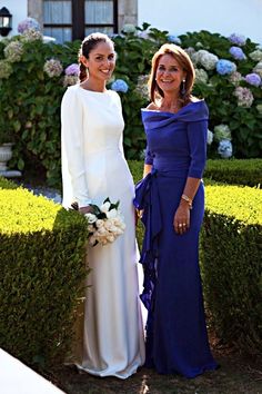 two women standing next to each other in front of some bushes and flowers on the ground