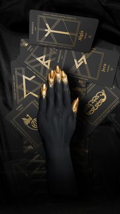 A black and gold painted hand, above some cards from The Grid of Fortune Rune Oracle Deck. The runes Algiz, Berkana, Jera, Pertro are visible and the animal fylgja cards The Serpent and The Owl. White Magic