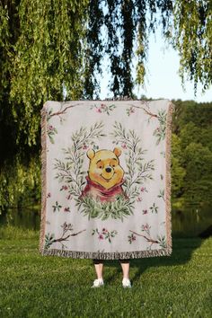 a woman holding up a winnie the pooh blanket in front of a tree and water