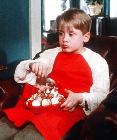 a young boy sitting in a chair eating food from a bowl on his lap and looking at the camera