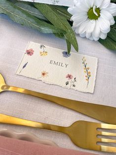 a place setting with gold utensils, flowers and a name tag on it