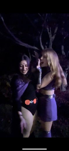 two women dressed up in costumes standing next to each other at night with trees behind them