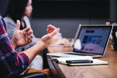 two people sitting at a table with laptops in front of them and one person holding their hands up