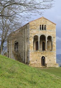 an old stone building with arched windows on top of a grassy hill next to trees