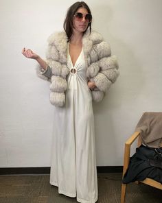 a woman standing in front of a wall wearing a white dress and large fur coat