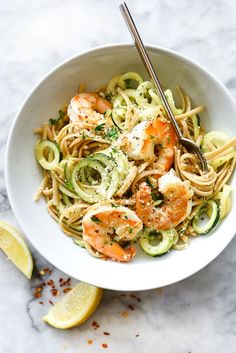 a white bowl filled with pasta and shrimp on top of a marble table next to lemon wedges