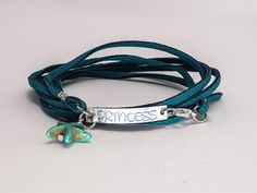 "Hand-stamped wrap bracelet for a favorite princess. Fitted as a 7\" to a 7-1/2\" bracelet, it adjusts smaller or larger depending on how many loops you make. Bracelet is a long strand of teal faux suede--so the color will not bleed--with a stamped aluminum plate with blue lettering. Lobster claw clasp makes it easy to latch. Coordinating necklace is also available. Choose between Pinafore or Fantasy lettering. Not sure how to use it? See a video of how to wear the bracelet at https://youtu.be/B Handmade Adjustable Green Name Bracelet, Adjustable Personalized Turquoise Friendship Bracelets, Adjustable Nickel-free Name Bracelet, Adjustable Nickel Free Name Bracelet, Adjustable Wrap Bracelet For Friendship, Nickel-free Adjustable Bangle Wrap Bracelet, Adjustable Hand Stamped Bracelets, Adjustable Hand Stamped Bohemian Bracelets, Adjustable Hand Stamped Bracelet