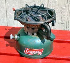 an old gas stove sitting on top of a red wooden bench with the word coleman's written on it