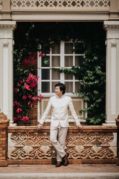 a man in a white suit and tie standing on a balcony next to pink flowers