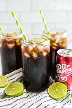 three glasses filled with soda and limes on top of a striped towel next to two cans