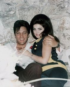 a man sitting next to a woman on top of a bed in front of a stone wall