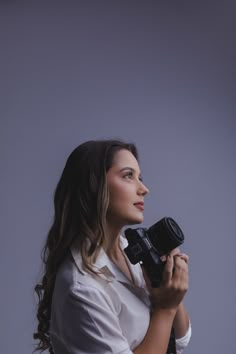 a woman holding a camera in her right hand and looking away from the camera, against a gray background