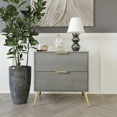 a grey and gold chest of drawers next to a plant in a gray vase with a white lamp on top