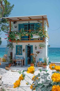 a white house with blue shutters and yellow flowers on the front porch next to the ocean